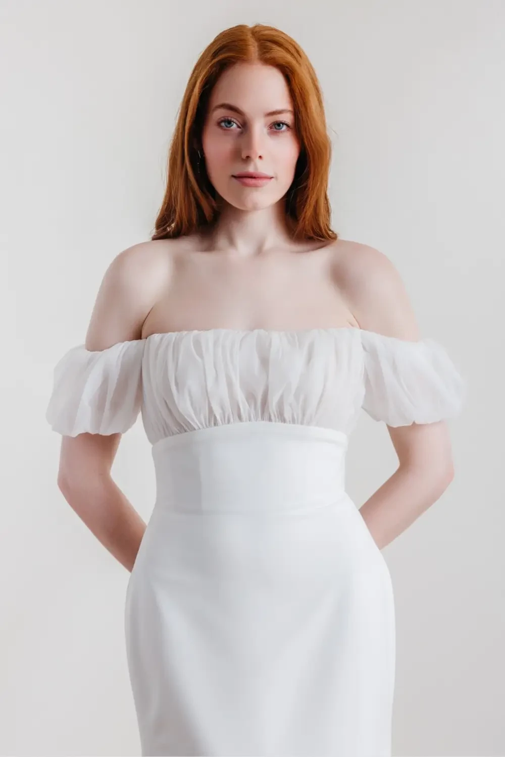 A woman in a white off-shoulder dress with puffed sleeves, standing with her hands at her sides against a plain background.
