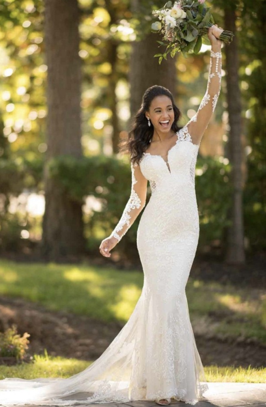 Model is wearing a long sleeve lace sheath wedding gown with sheer side cutouts, a deep plunging neckline, a dramatic cathedral train, and a fully open lace-trimmed back.