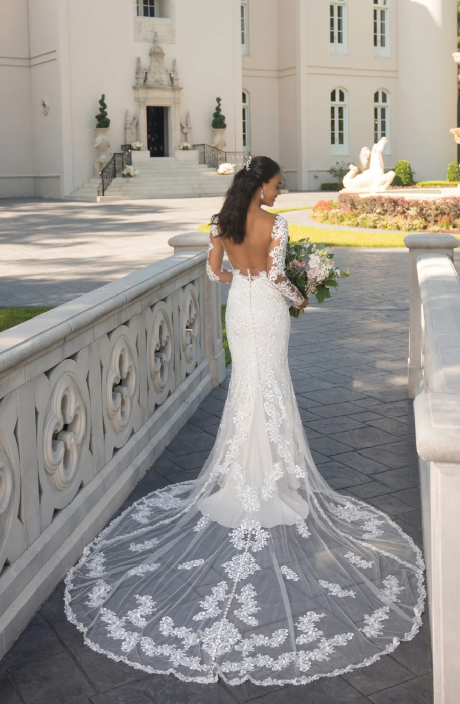 Model is wearing a long sleeve lace sheath wedding gown with sheer side cutouts, a deep plunging neckline, a dramatic cathedral train, and a fully open lace-trimmed back.