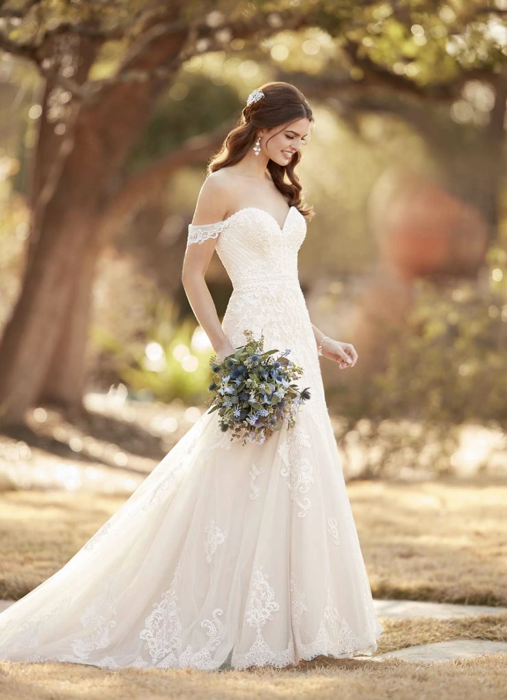A woman in a white off-the-shoulder wedding dress standing outside, holding a bouquet of flowers, with trees and soft lighting in the background.