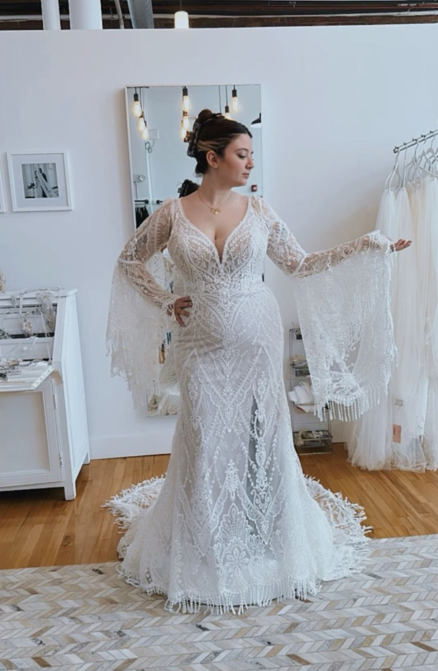 A woman in a detailed white lace wedding gown with bell sleeves stands confidently in a chic, bright studio. The atmosphere is elegant and poised.