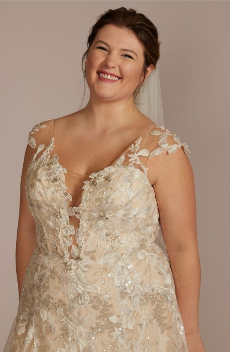Smiling bride in an embellished, floral lace wedding dress with sheer sleeves against a neutral background, conveying joy and elegance.