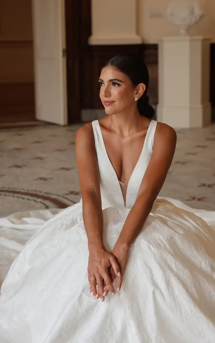 A bride wearing a sleeveless white wedding dress, sitting on the floor with her legs crossed, looking off to the side.