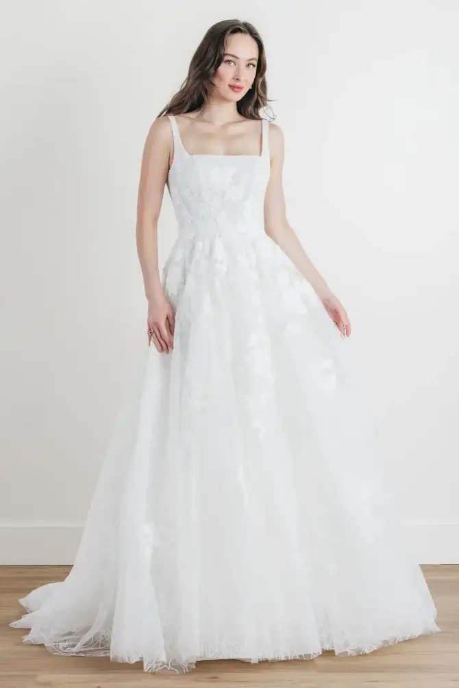 A woman in a white wedding dress with a square neckline, standing against a plain background.