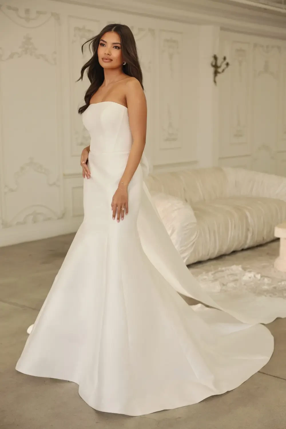 A woman in a strapless white gown standing elegantly in a stylish interior.