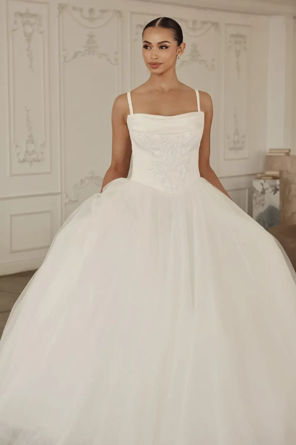 A model wearing a white wedding gown with a fitted bodice and a full tulle skirt, standing in an elegantly decorated room.