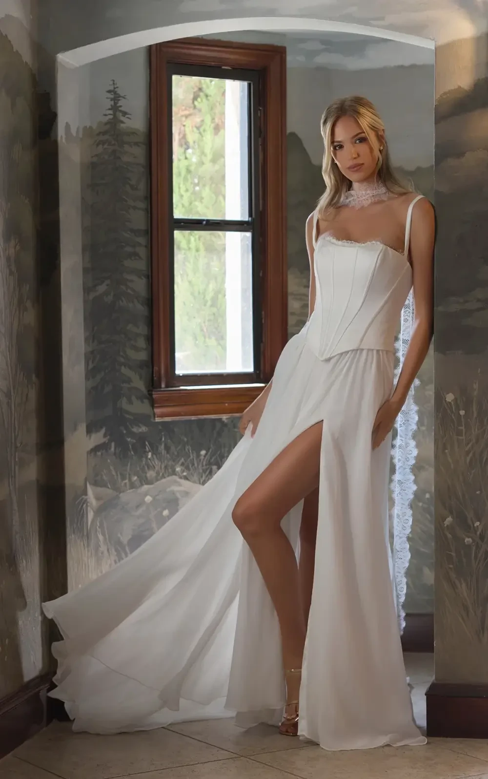 A model wearing a white gown with a high slit stands in a room with painted walls and a window.