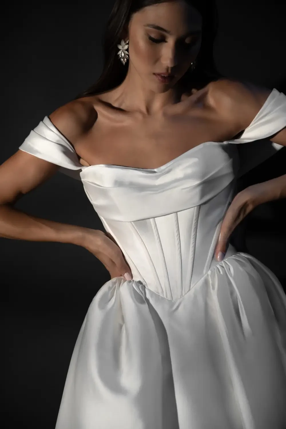 A woman wearing an elegant off-shoulder white dress, featuring a fitted bodice and a flowing skirt. She is posing with her hands on her waist and looking down.