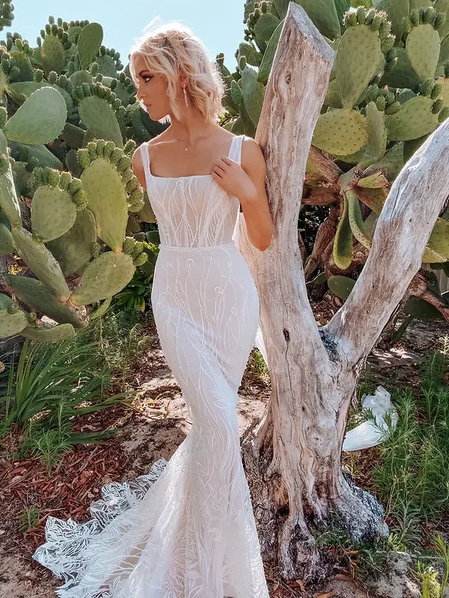 Model is wearing a curve-hugging lace fit-and-flare gown with a square neckline, sleeveless bodice, and low open square back.