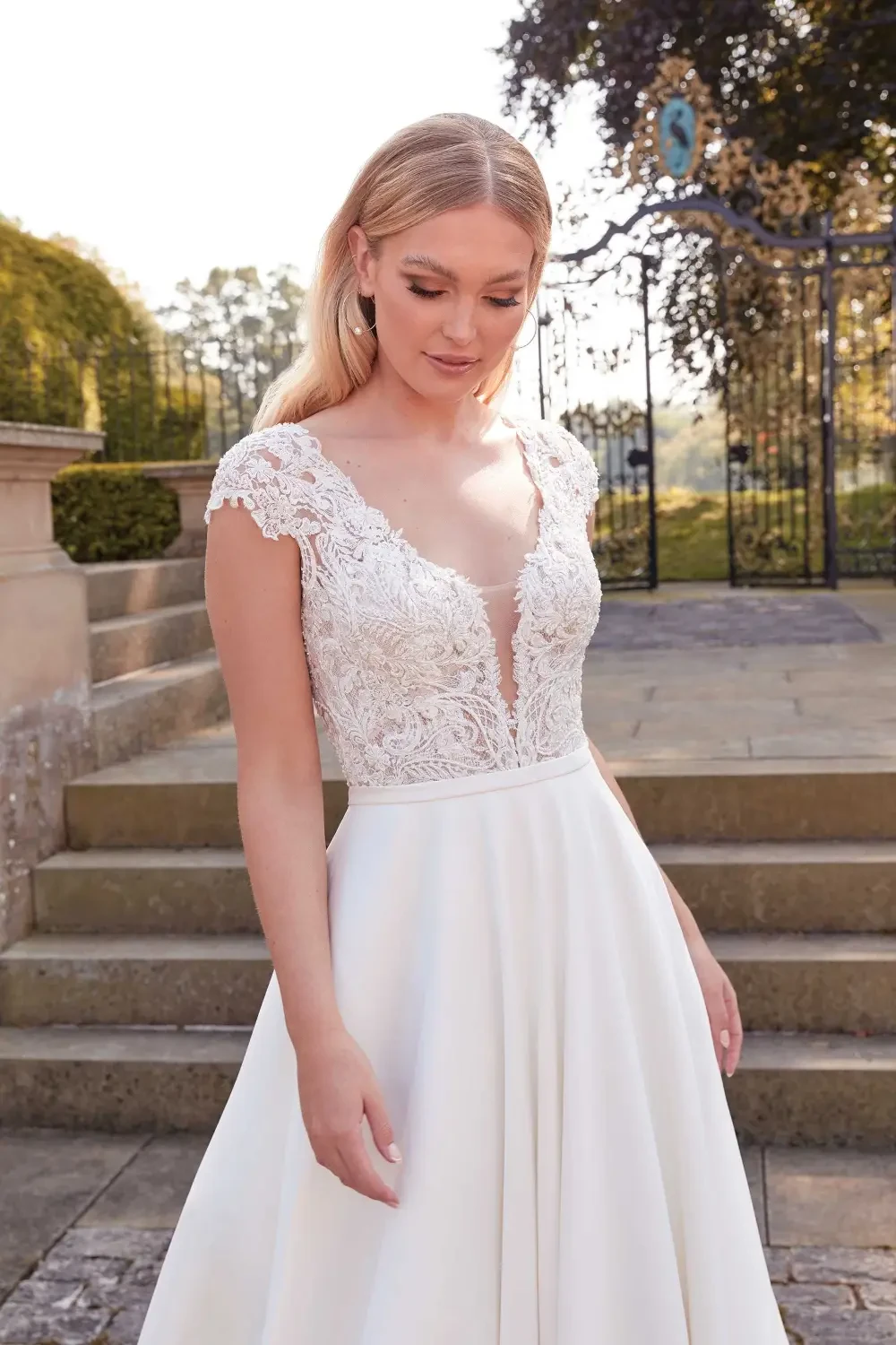A woman in a white wedding dress with a lace bodice and short sleeves, standing outside near a stone path and greenery.