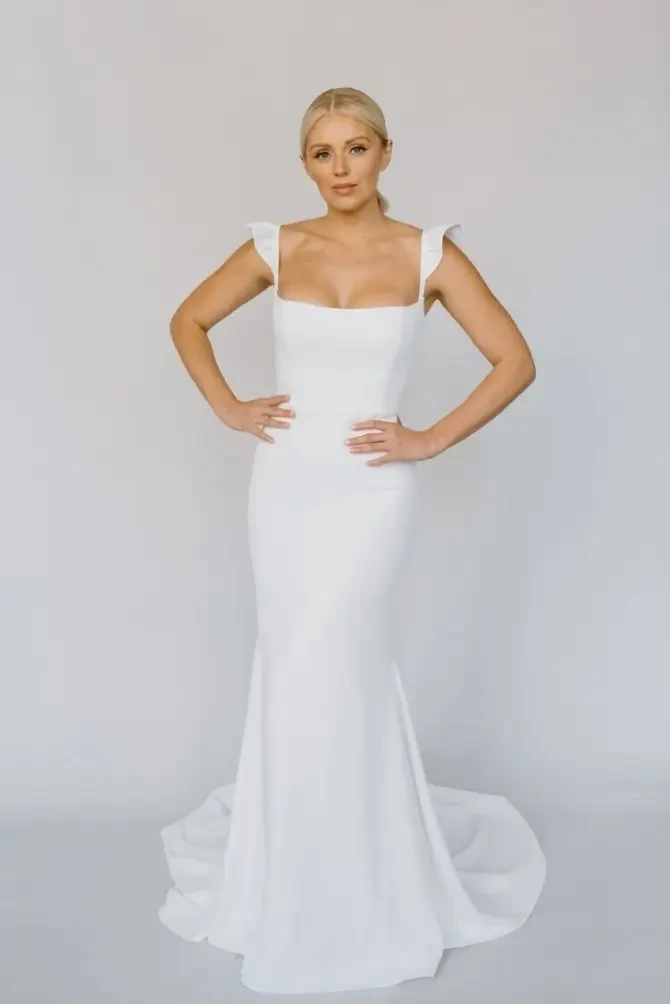 Woman in a fitted white gown stands confidently against a light gray background. The dress features ruffled shoulder details and a long, flowing skirt.
