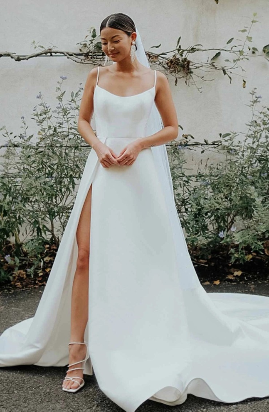 Bride in sleek white gown with a thigh-high slit, standing outdoors. Soft smile, veil draped, greenery and vine-covered wall in background. Elegant and serene.